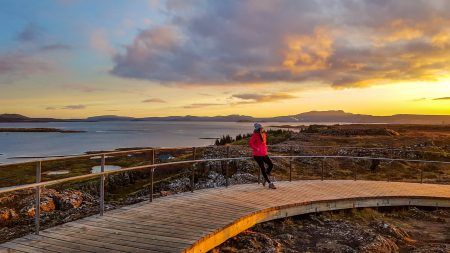 A beautiful girl wearing a pink jacket, leaning on a barrier during the sunset over national park Þingvellir in Iceland.