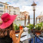 An elegant woman with a red hat enjoys an aperitif sitting next to the Grand Canal in Venice, Italy
