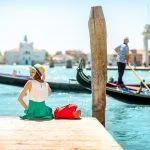 Young woman traveler sitting on the pier enjoying a beautiful view of the venetian chanal with gondolas floating in venice