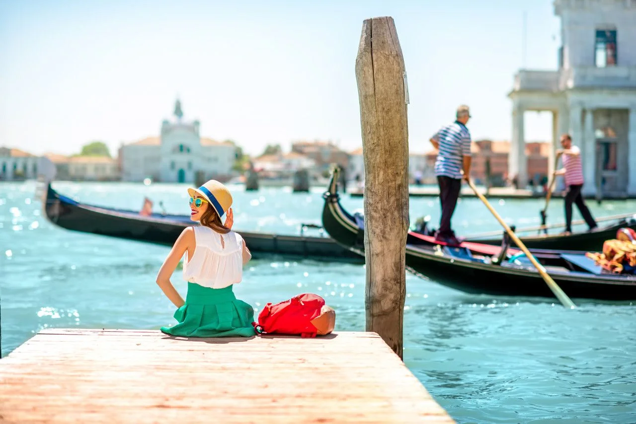 Young woman traveler sitting on the pier enjoying a beautiful view of the venetian chanal with gondolas floating in venice