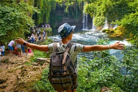 A woman stands with outstretched arms near a waterfall. Tourist with a backpack on the background of a waterfall and rainforest. The Upper Duden Waterfall