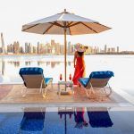 Beautiful beach in Dubai with a lady in red and white hat, 2 sun chairs and an umbrella. In the background is the skyline of Dubai Marina