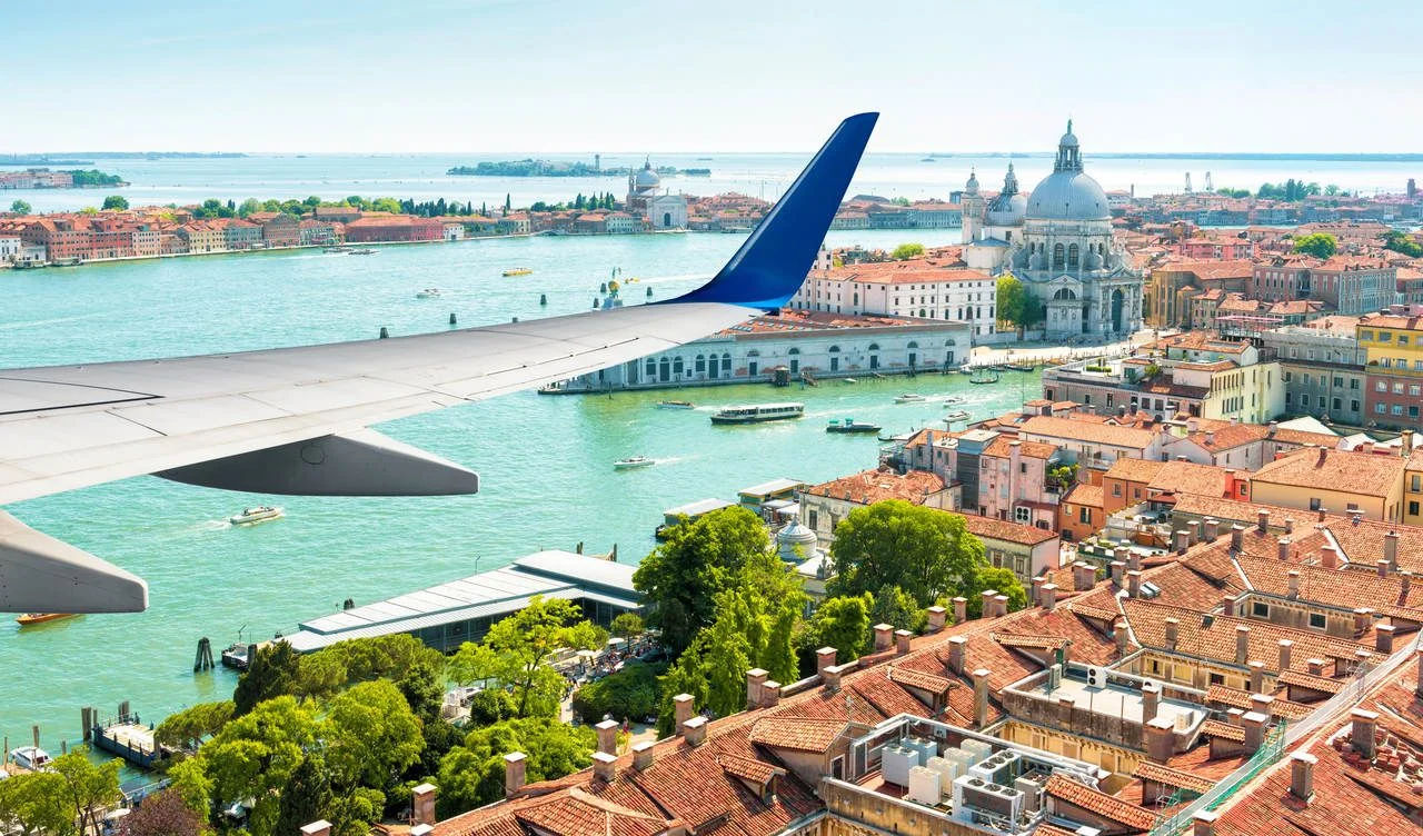 Plane flies above Venice, Italy. Aerial panoramic view of city and sea from airplane window. The plane’s wing over Venice during taking off or landing. Concept of flight, vacation and summer travel.