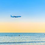 Airplane flying low above sea and people tourists swimming in water, clear blue orange sky at sunset, plane preparing to land at Larnaca airport above Mediterranean sea