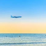 Airplane flying low above sea and people tourists swimming in water, clear blue orange sky at sunset, plane preparing to land at Larnaca airport above Mediterranean sea