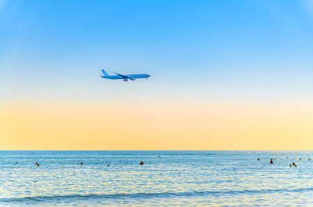 Airplane flying low above sea and people tourists swimming in water, clear blue orange sky at sunset, plane preparing to land at Larnaca airport above Mediterranean sea