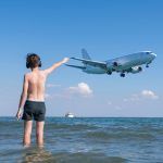 Teen boy in the wave of the sea on the beach in the headphones under the landing planes. Traveling on an airplane with children.