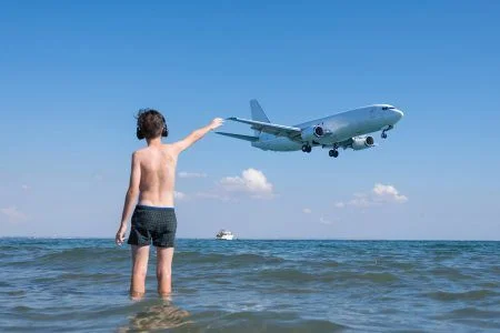 Teen boy in the wave of the sea on the beach in the headphones under the landing planes. Traveling on an airplane with children.