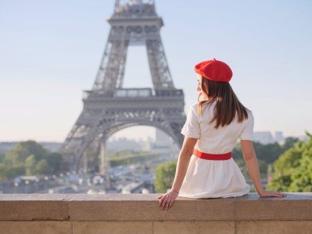 A beautiful girl in a white dress, a red beret sits against the backdrop of the Eiffel Tower in Paris. Travel to Paris. Ideas for a photo shoot.