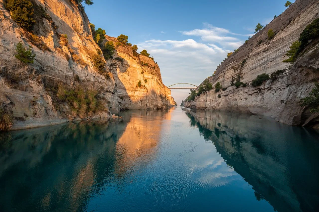Seascape of both sides of Corinth’s canal coast, left seaside glows in a golden sunrise light, right is in shadow, bridge contour and blue skies on background, noisy blurred water reflcetion
