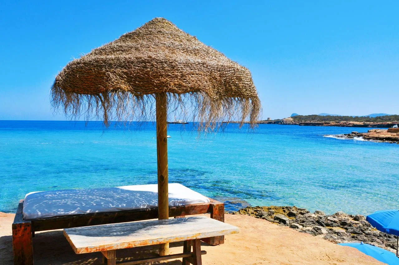 detail of a relaxing area in a Cala Conta beach in Ibiza Island, Spain, with a comfortable sunlounger and a rustic umbrella made of natural fibers