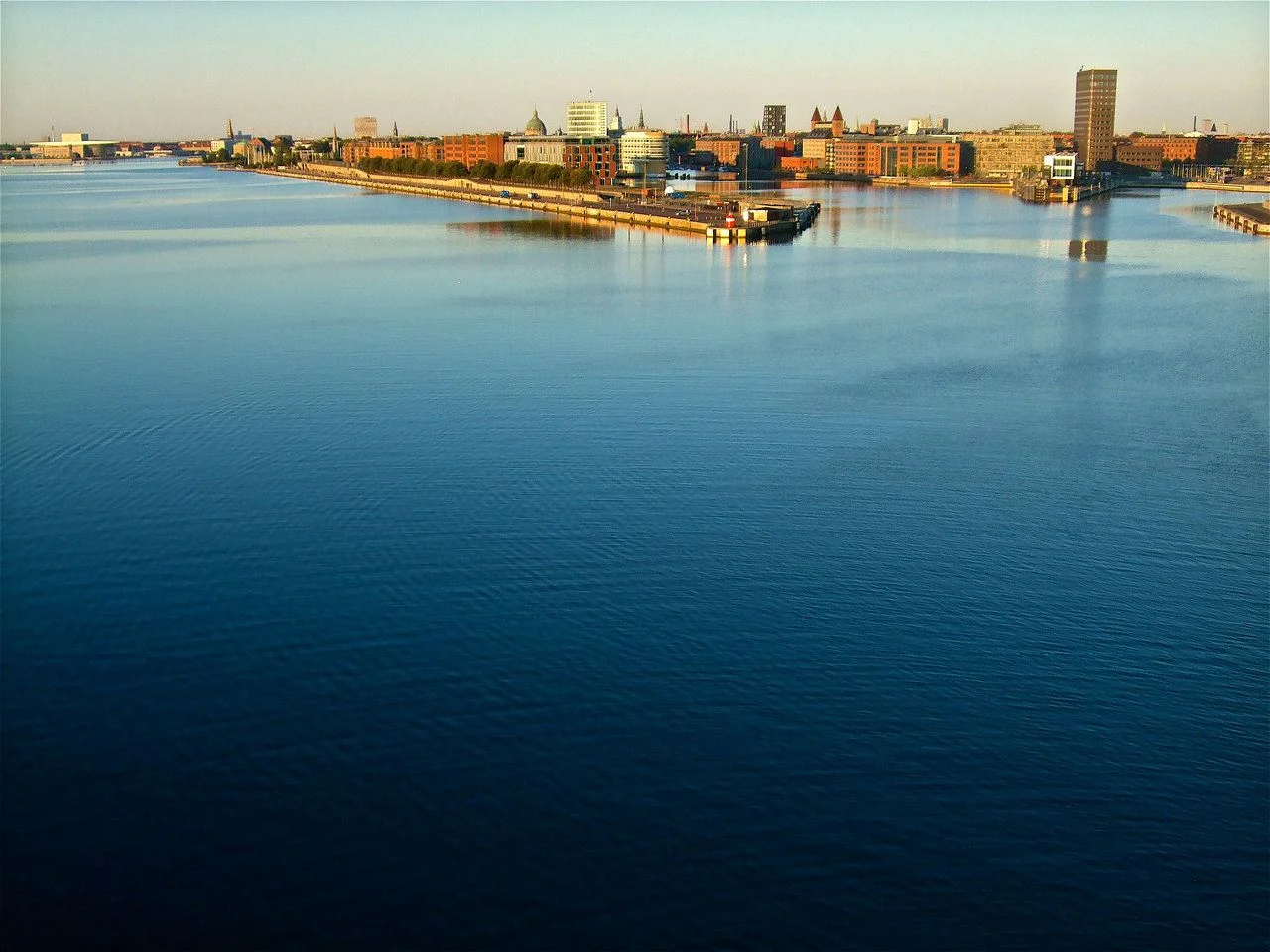 Morning in Copenhagen from the cruise ship terminal, with a view of the city on the horizon, the opera house, spires and domes of several landmarks prominent, in tones of blue, orange and tan