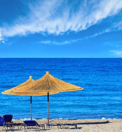 Summer morning pebbly beach with sunbeds and strawy sunshades (Borsh, Albania). Blue sky with some clouds.