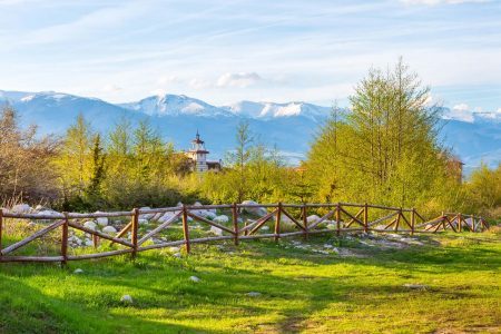 Bansko, Bulgaria spring landscape with the wooden fence, trees, tower of chalet and snowy Rila mountains peaks