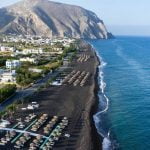 Top view of Perissa beach on the Greek island of Santorini with sunbeds and umbrellas. Beach is covered with fine black sand, and drops off sharply into the water.