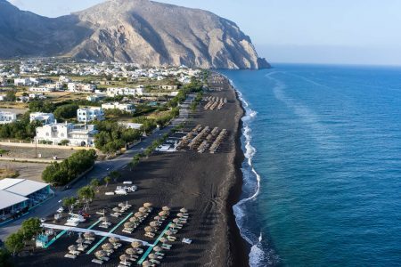 Top view of Perissa beach on the Greek island of Santorini with sunbeds and umbrellas. Beach is covered with fine black sand, and drops off sharply into the water.